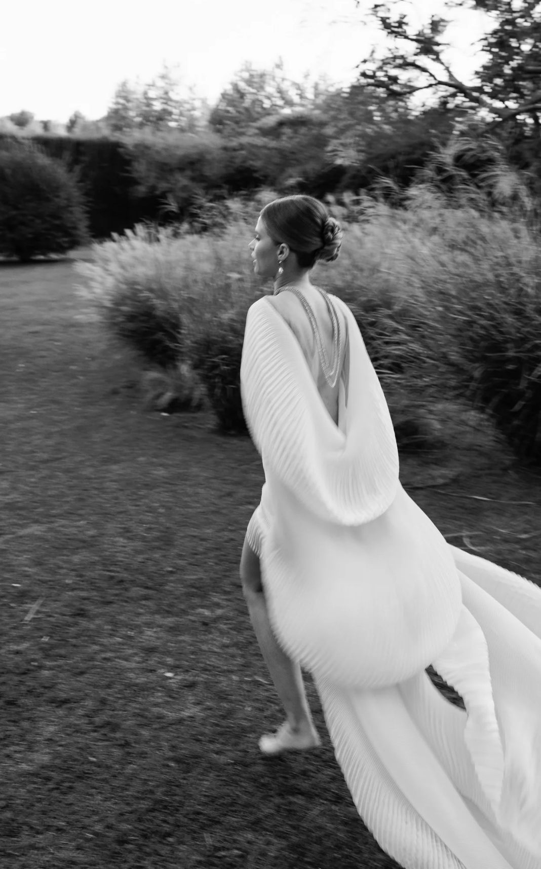 A woman dressed in a flowing, elegant gown walking outdoors in a natural setting with trees and bushes in the background.