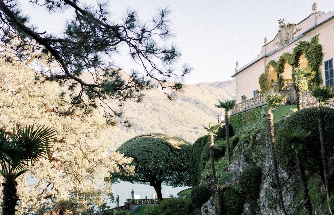 A scenic view of a luxurious estate with a pastel-colored mansion, lush greenery, palm trees, and a lake in the background, with mountains in the distance.