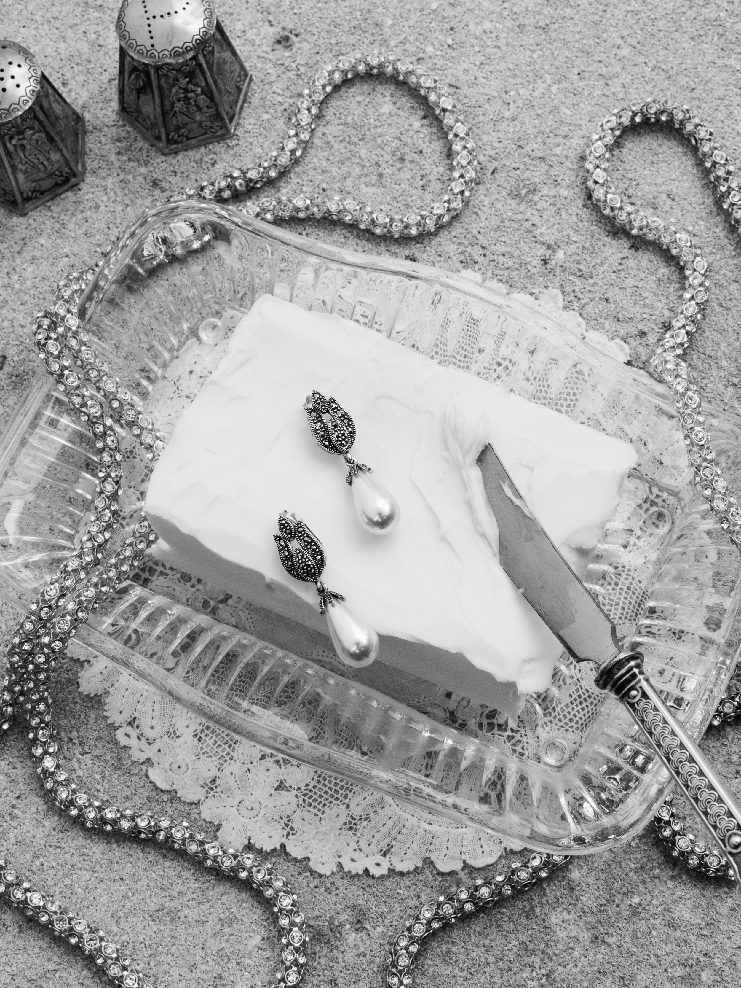 A glass serving dish with a block of white cake, decorated with dangling pearl earrings, a cake knife, and surrounded by rhinestone jewelry and two decorative jars on a lace doily.