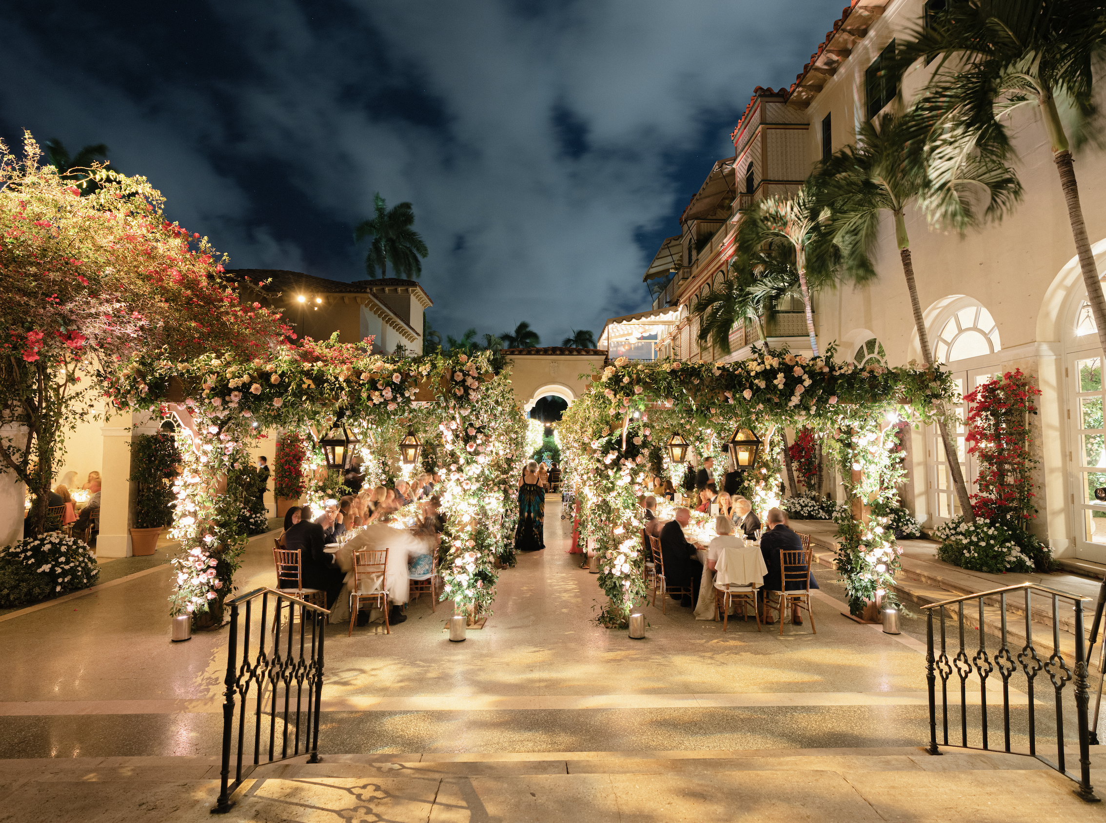 Nighttime outdoor event with tables and chairs under floral arches illuminated by lanterns and string lights, with guests dining in a courtyard surrounded by palm trees and elegant buildings.