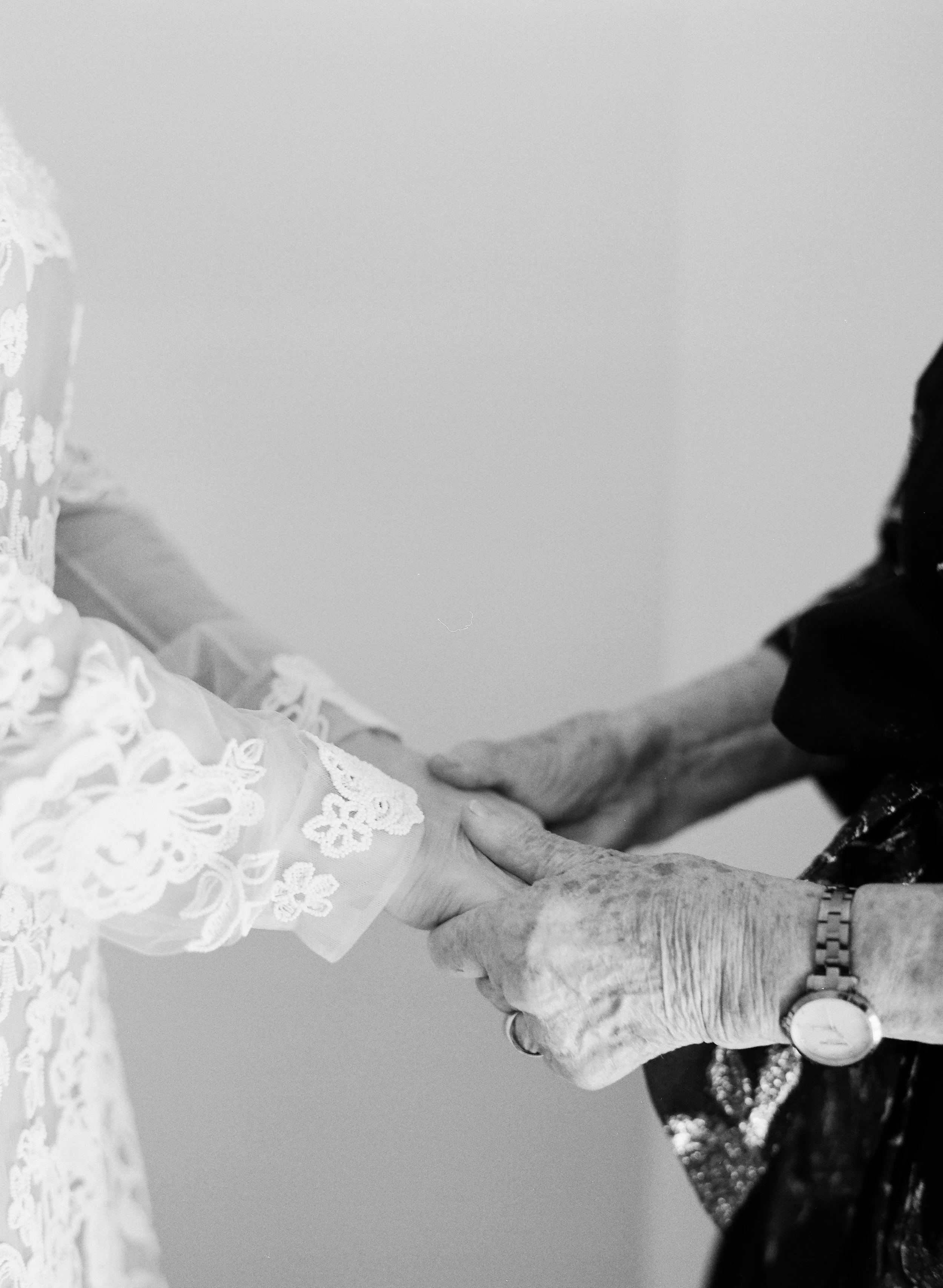 Close-up of two elderly women holding hands, one wearing a lace dress and the other wearing a dark outfit, in an intimate moment.