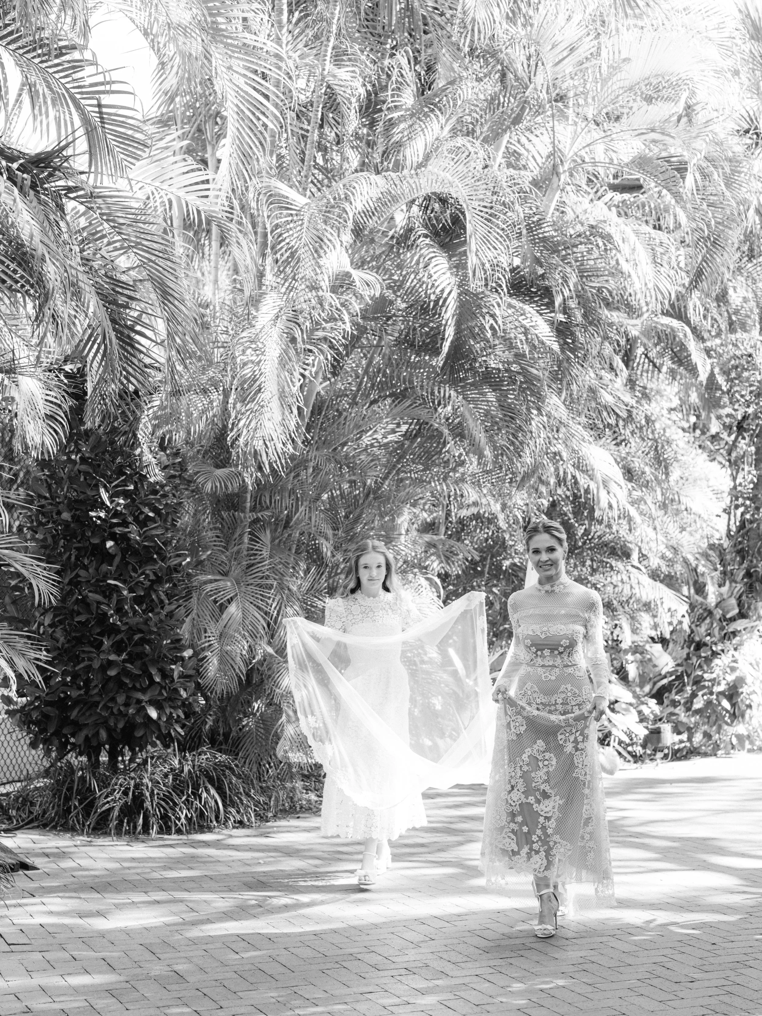 Two women dressed in elegant lace dresses walking on a brick path surrounded by lush tropical plants. One woman is holding a sheer fabric, and both are smiling.