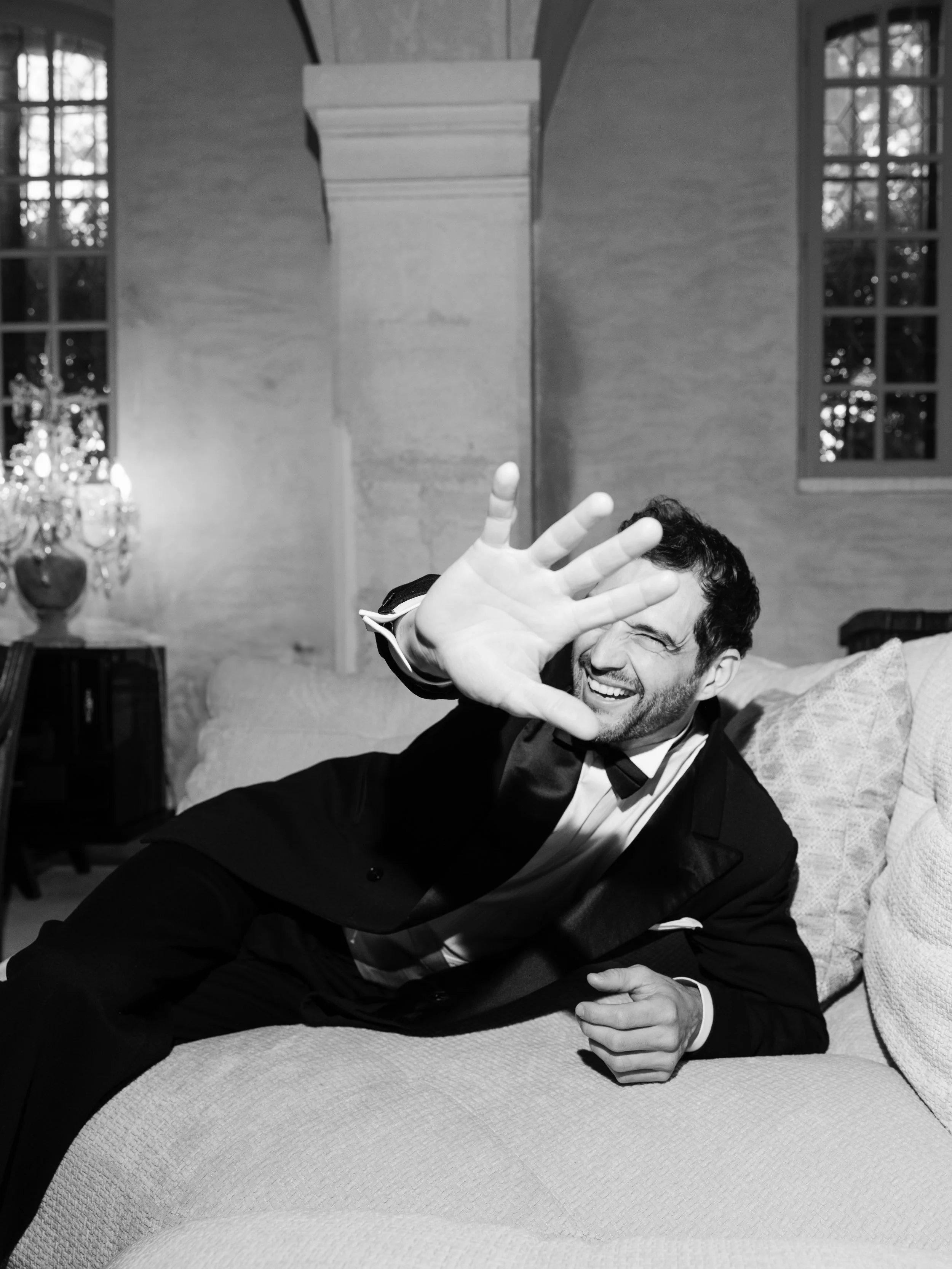 A man in a tuxedo is lying on a sofa, smiling and covering his face with his hand.