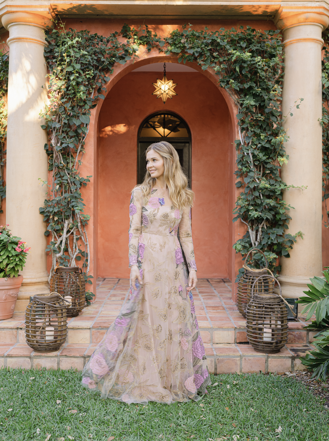 A woman in a long, floral dress with purple and gold patterns standing on a brick patio in front of a pink stucco house with columns and green ivy. There's warm lighting hanging above her and wicker lanterns on the porch.