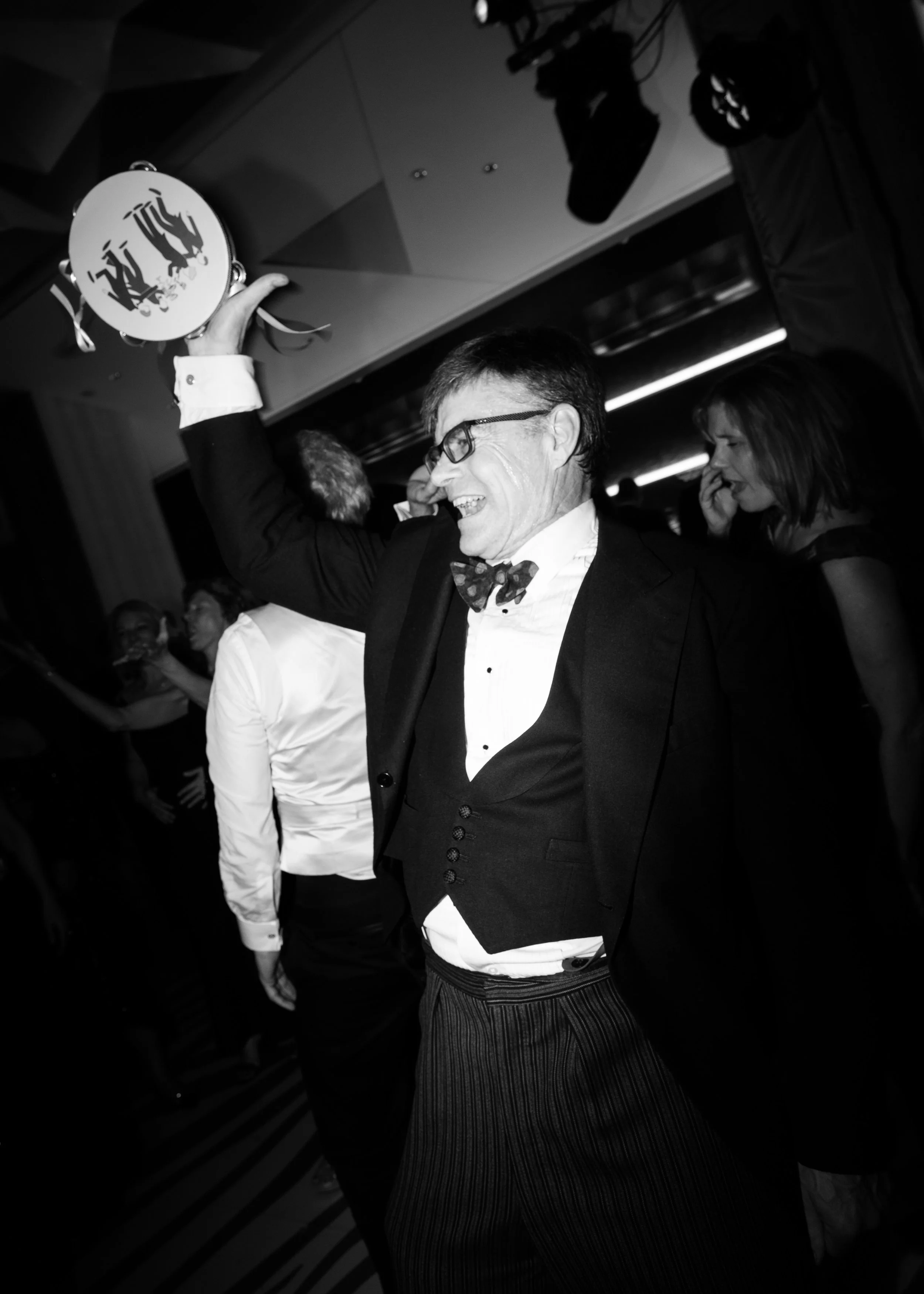 A man in formal attire, wearing a tuxedo and bow tie, is celebrating at a party, holding a tambourine overhead, with other people dancing and enjoying the event in the background.