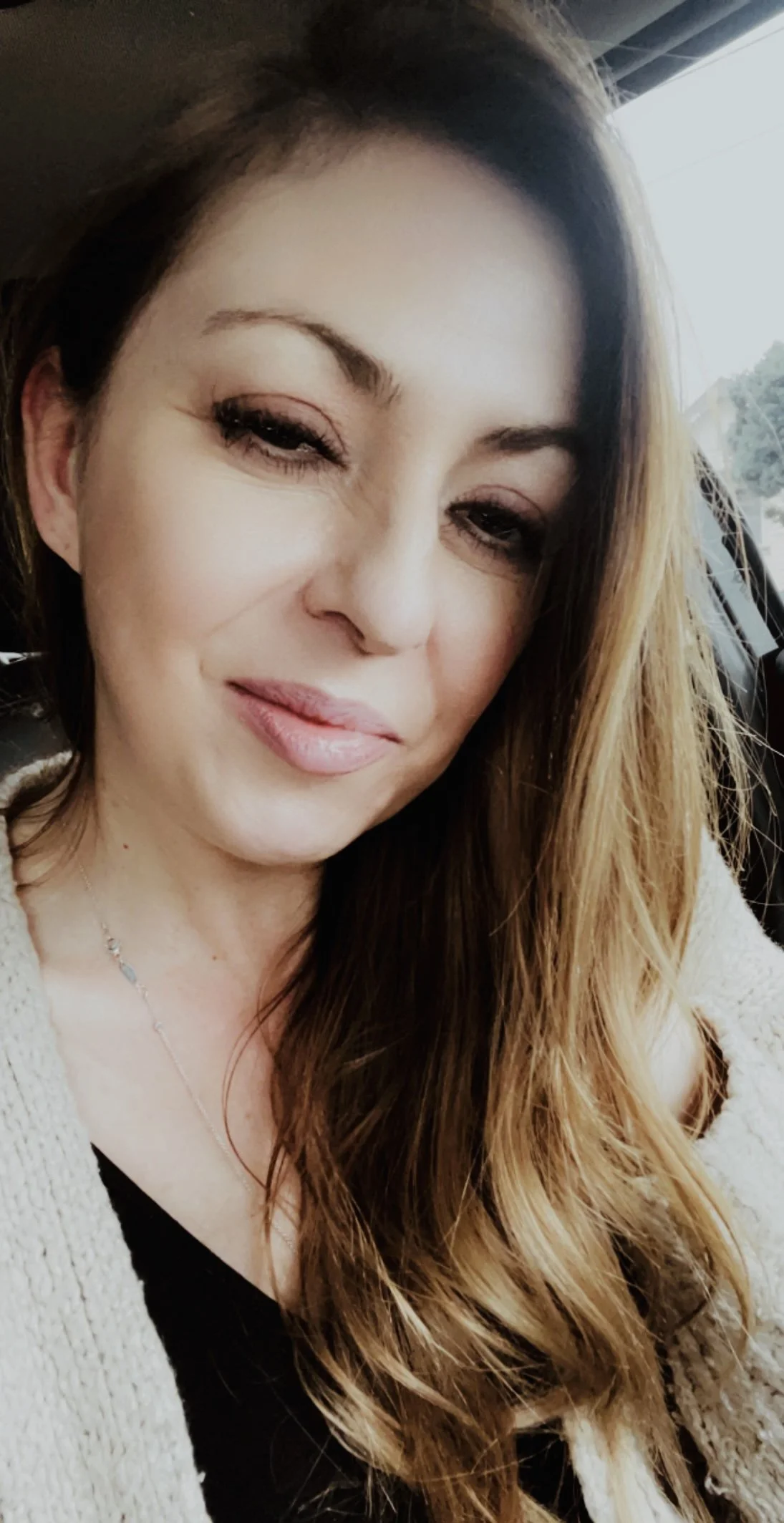 A woman with long brown hair, closed eyes, and a slight smile, sitting in a car.