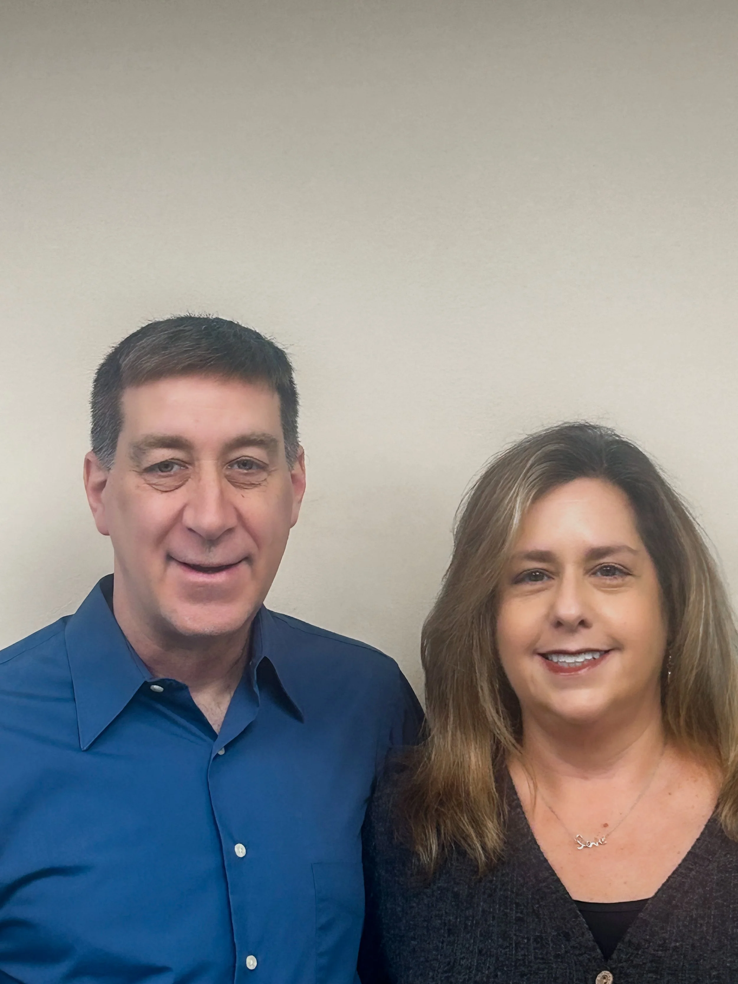 A man and a woman standing side by side against a plain wall, smiling at the camera.