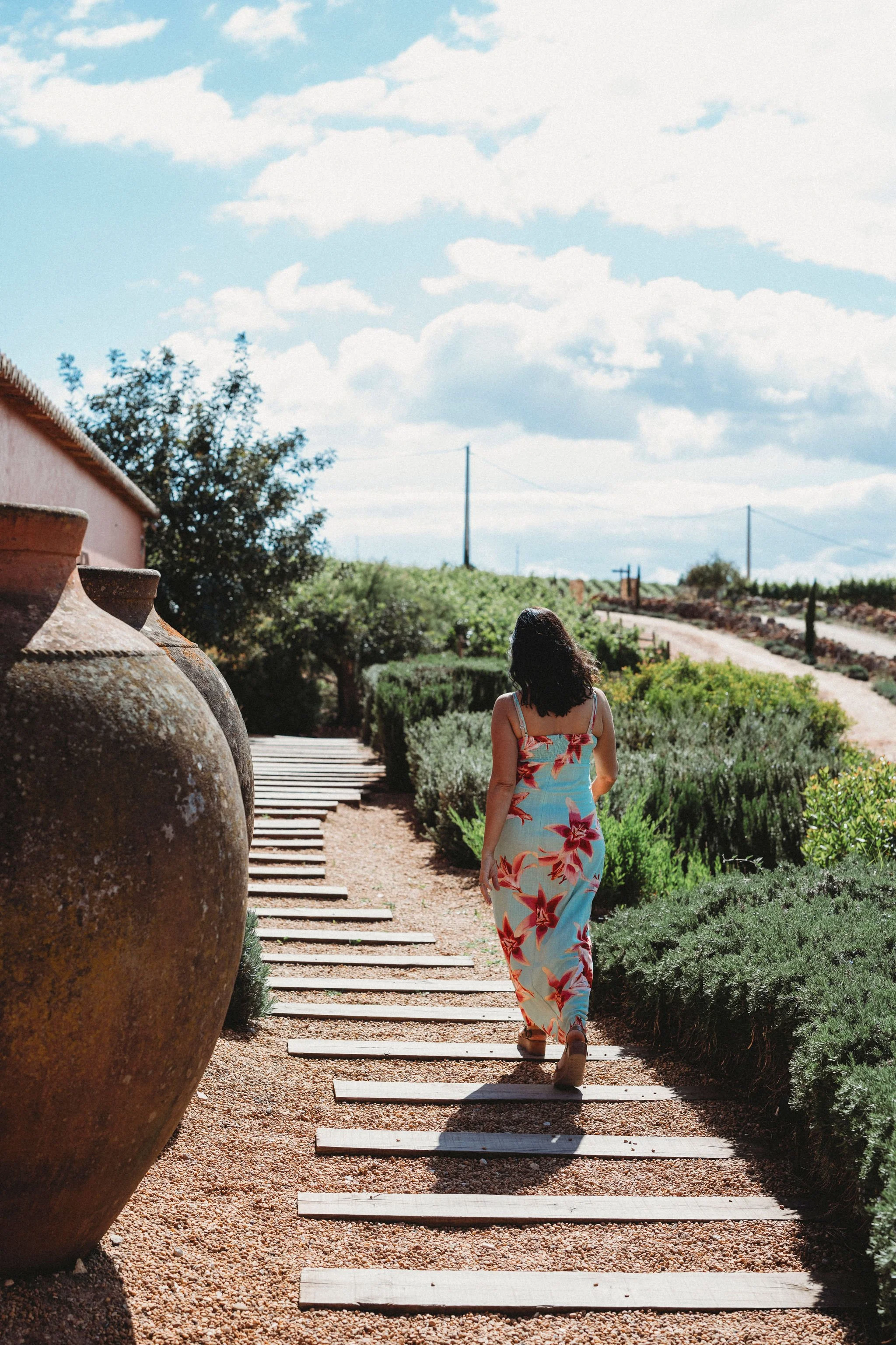 A woman in a floral dress walking along a wooden pathway through a garden with large clay pots and greenery, under a partly cloudy sky.