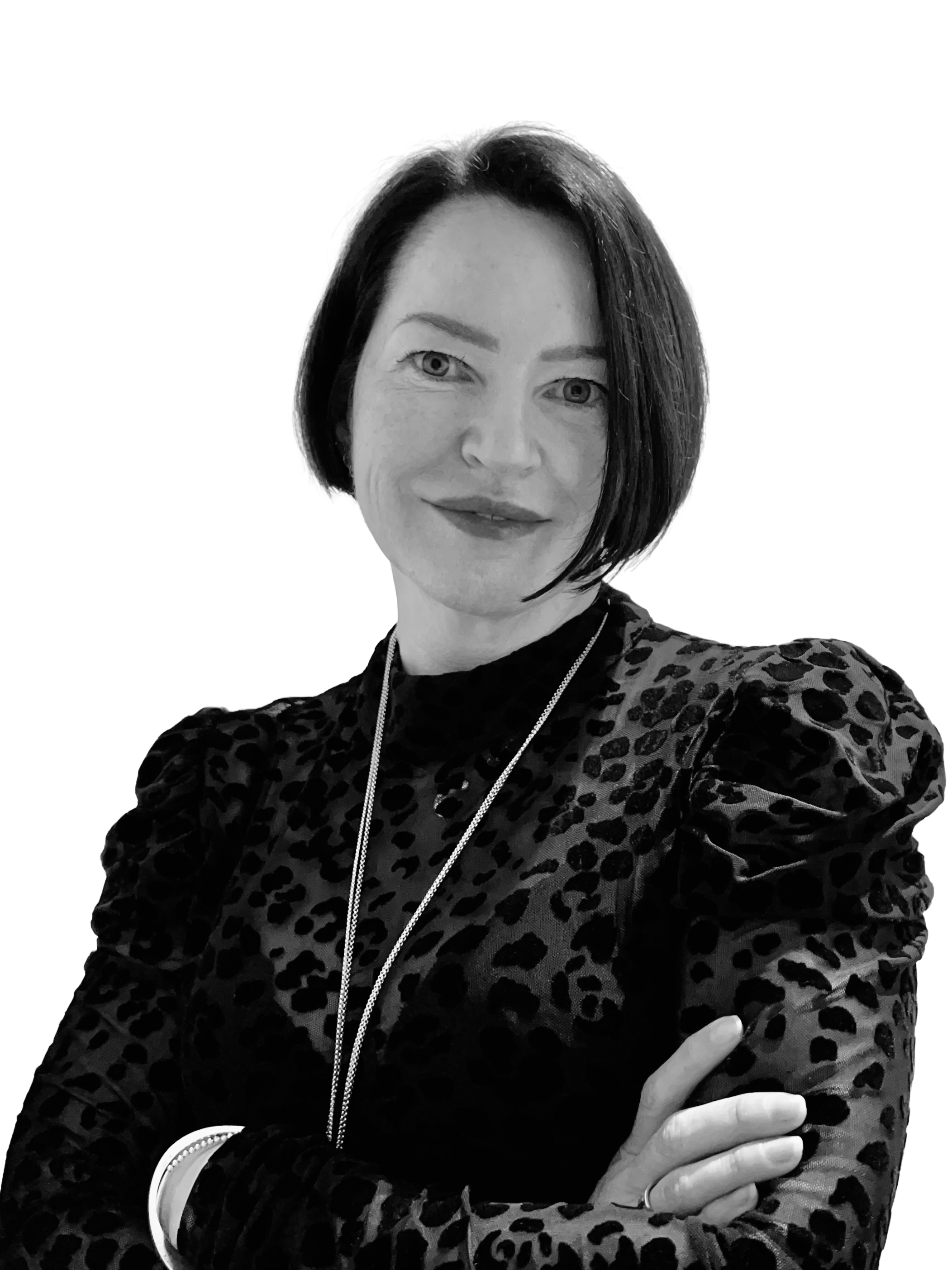 Black and white portrait of a woman with short dark hair, wearing a leopard print shirt and a long necklace, standing against a plain background with her arms crossed.