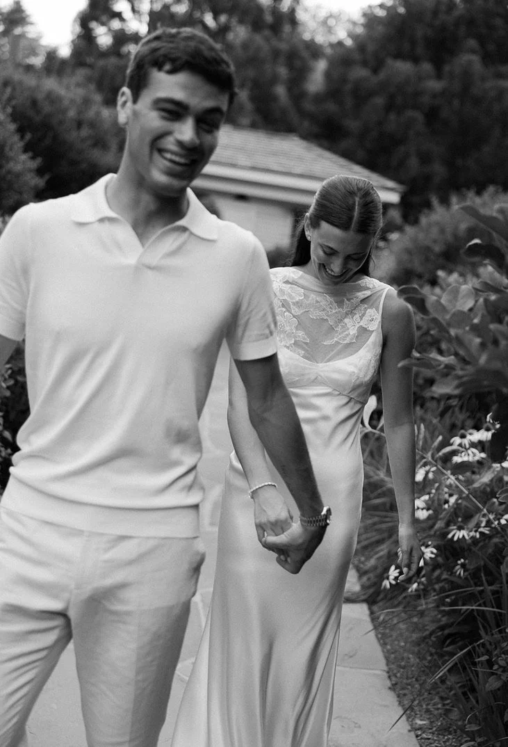 A black and white photo of a couple walking hand in hand outdoors, smiling and looking down. The man is wearing a polo shirt and light-colored pants, and the woman is in a satin dress with lace details, surrounded by plants and a house in the backgro