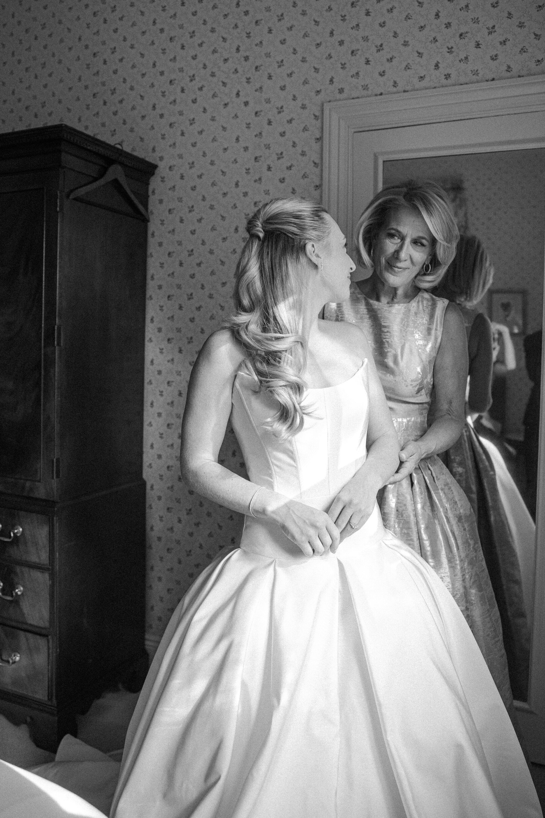 A bride in a wedding dress looking at her mother, who stands behind her and smiles, in a room with patterned wallpaper and a mirror reflecting another person.