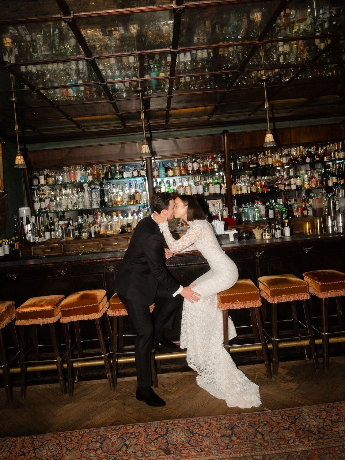 A bride and groom kissing at a bar, with the bride seated on the bar counter and the groom standing close to her, in a dimly lit venue with a large collection of liquor bottles behind them.