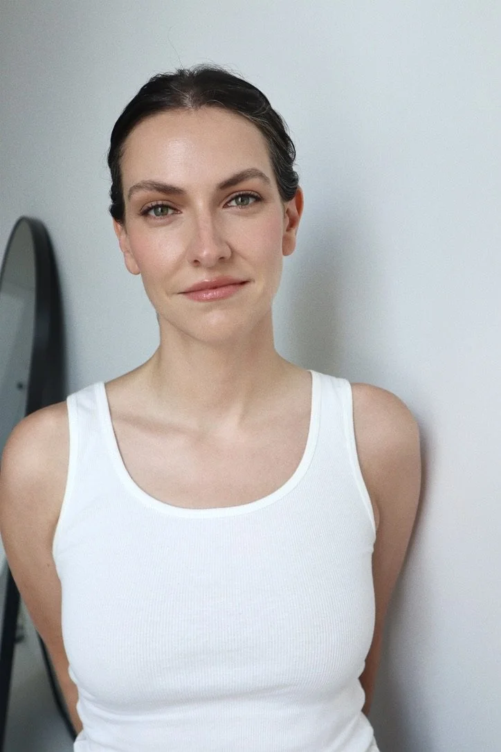 A young woman with dark hair in a braid, wearing a white sleeveless top, smiling slightly, standing in front of a plain light-colored wall.