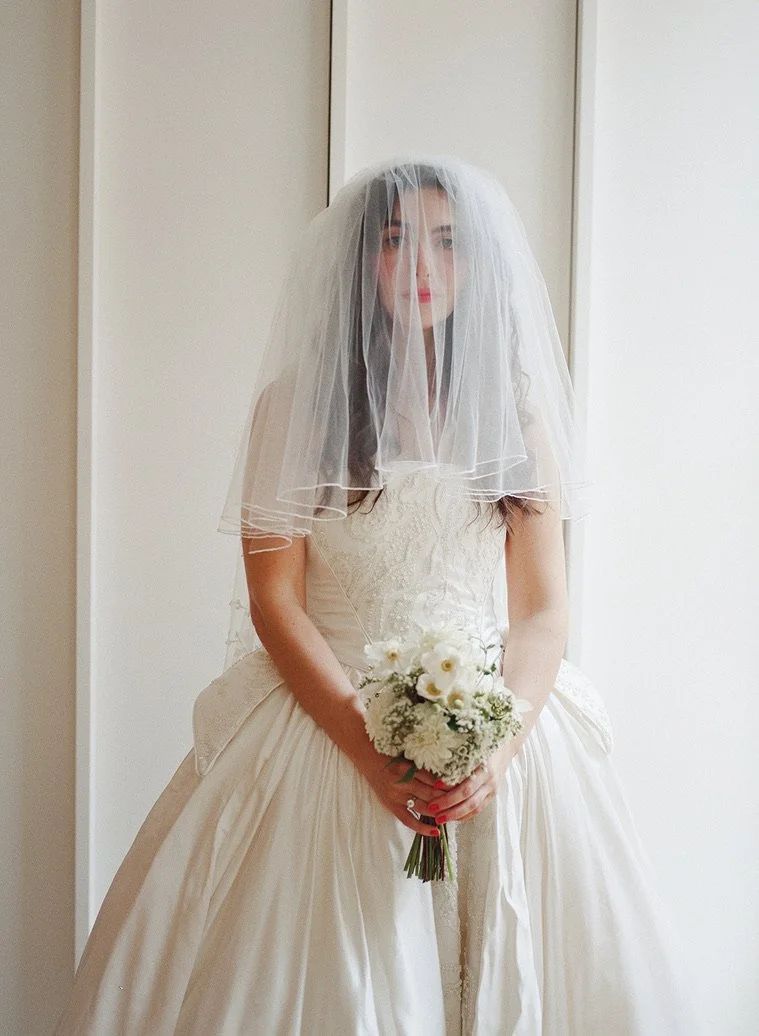 A bride in a white wedding gown holding a bouquet of white flowers, wearing a veil over her face, standing indoors with plain walls and a mirror behind her.