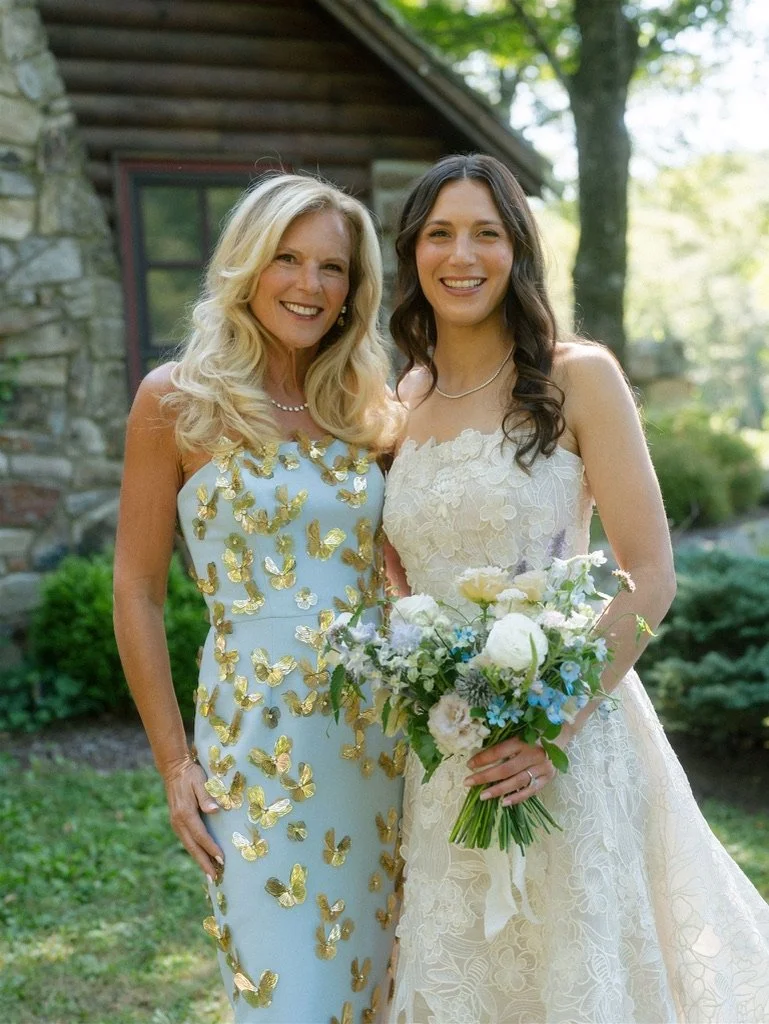Two women standing outdoors smiling, one in a white wedding dress holding a bouquet, and the other in a blue dress with gold butterfly patterns.