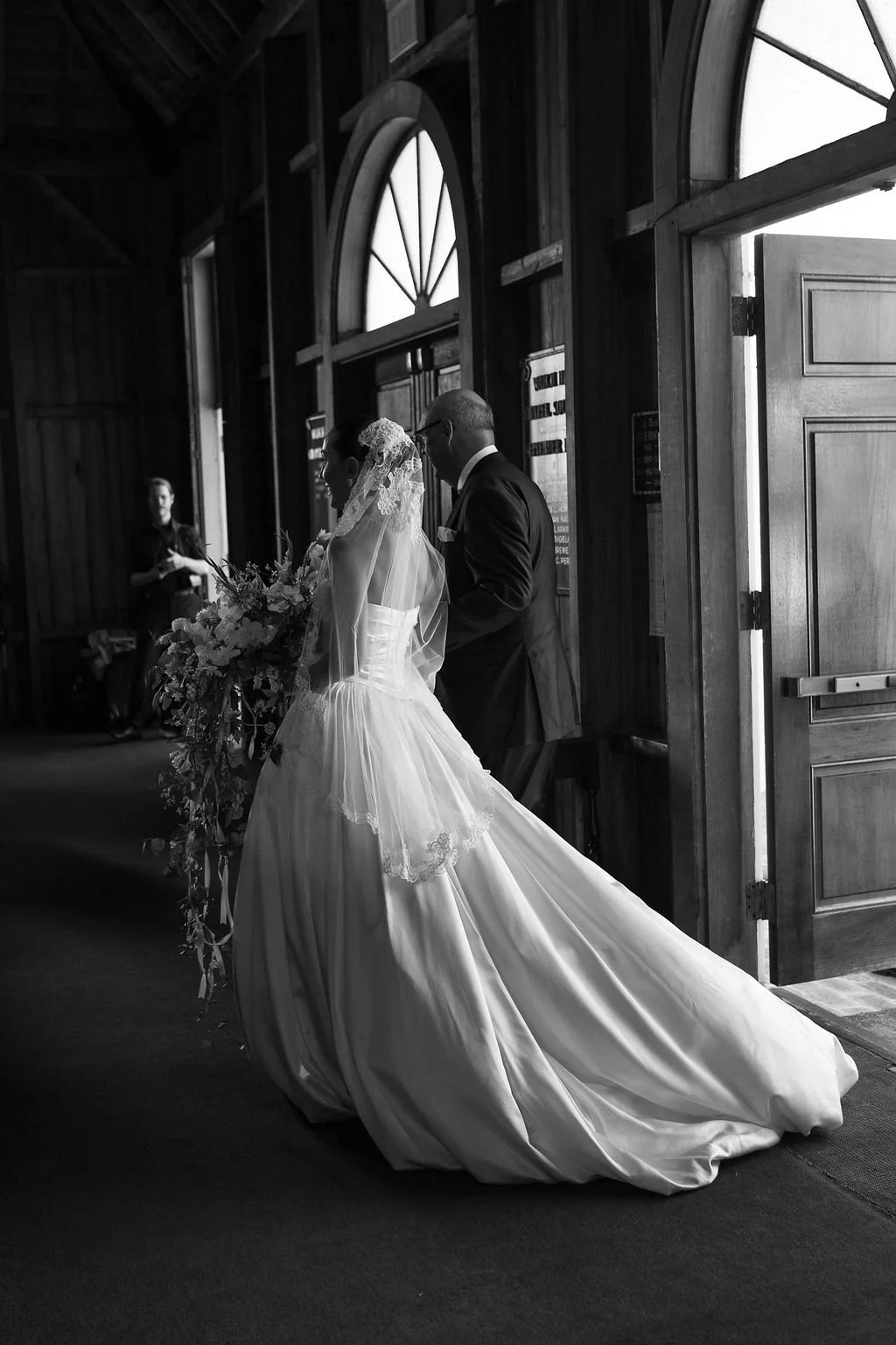 Black and white photo of a bride and groom standing in an indoor chapel, facing each other with the bride holding a bouquet, wooden walls, and large windows in the background.