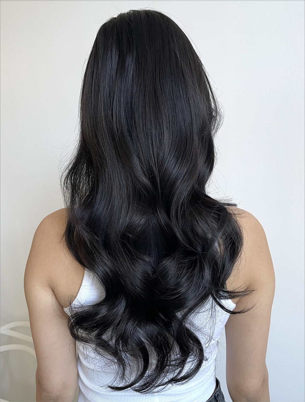 Back view of a woman with long, black, wavy hair over a white sleeveless top against a plain white background.