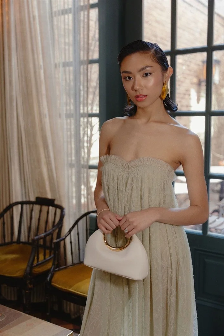 A woman with dark hair styled in waves, wearing a strapless beige dress with a textured pattern, standing indoors near large windows with a grid pattern. She has gold earrings and is holding a white handbag with a gold handle. In the background, ther