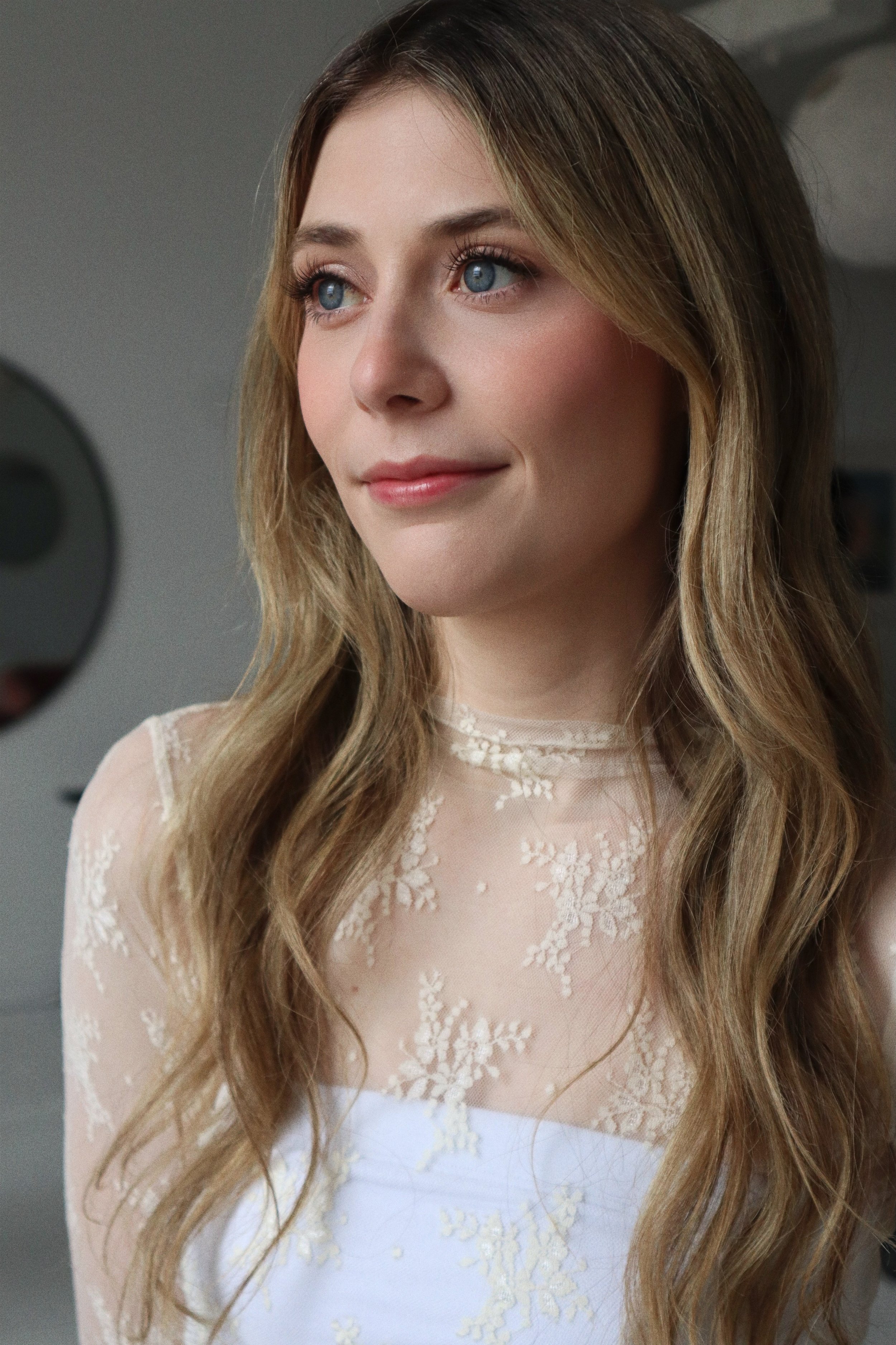 A young woman with long, wavy blonde hair and blue eyes, wearing a white dress with lace detailing.