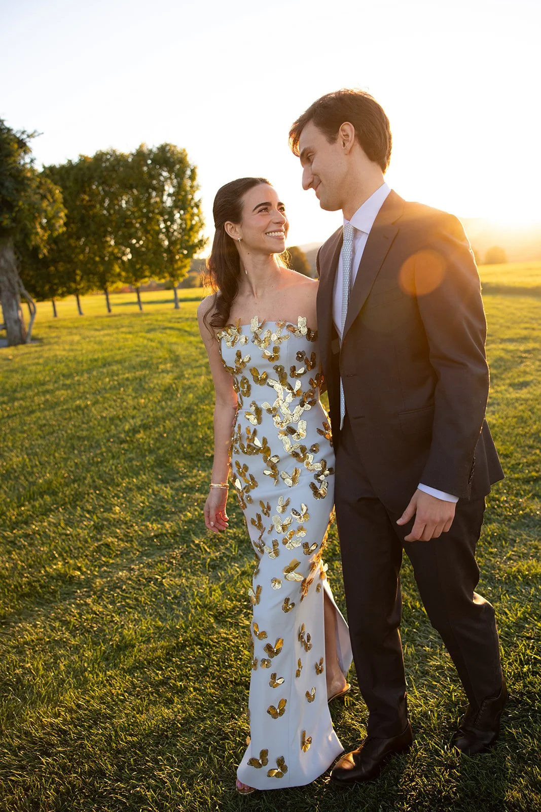 A couple dressed in formal attire walking in a grassy field during sunset, with the woman wearing a white Carolina Herrera gown adorned with gold butterflies and the man in a dark suit.