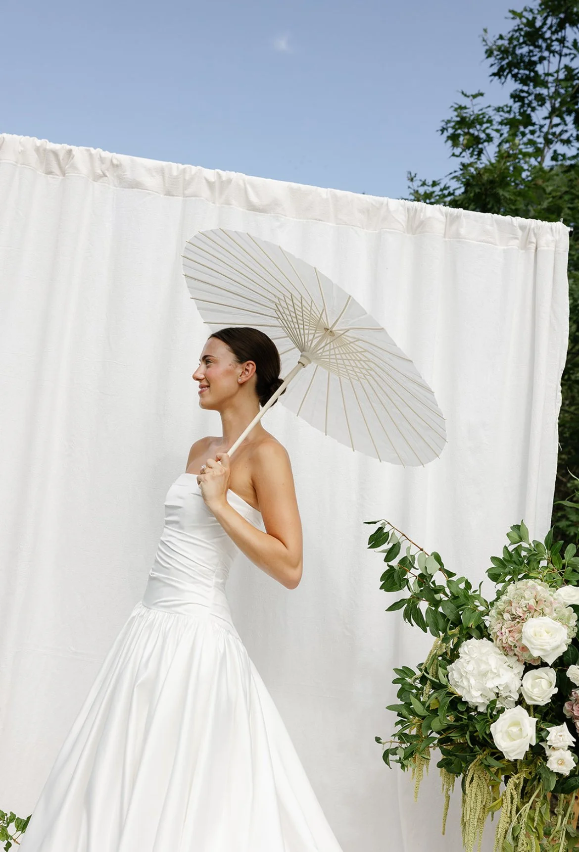 A woman in a white wedding dress holding a white parasol, standing beside a floral arrangement with white and pink flowers and greenery, outdoors with a white backdrop and blue sky.