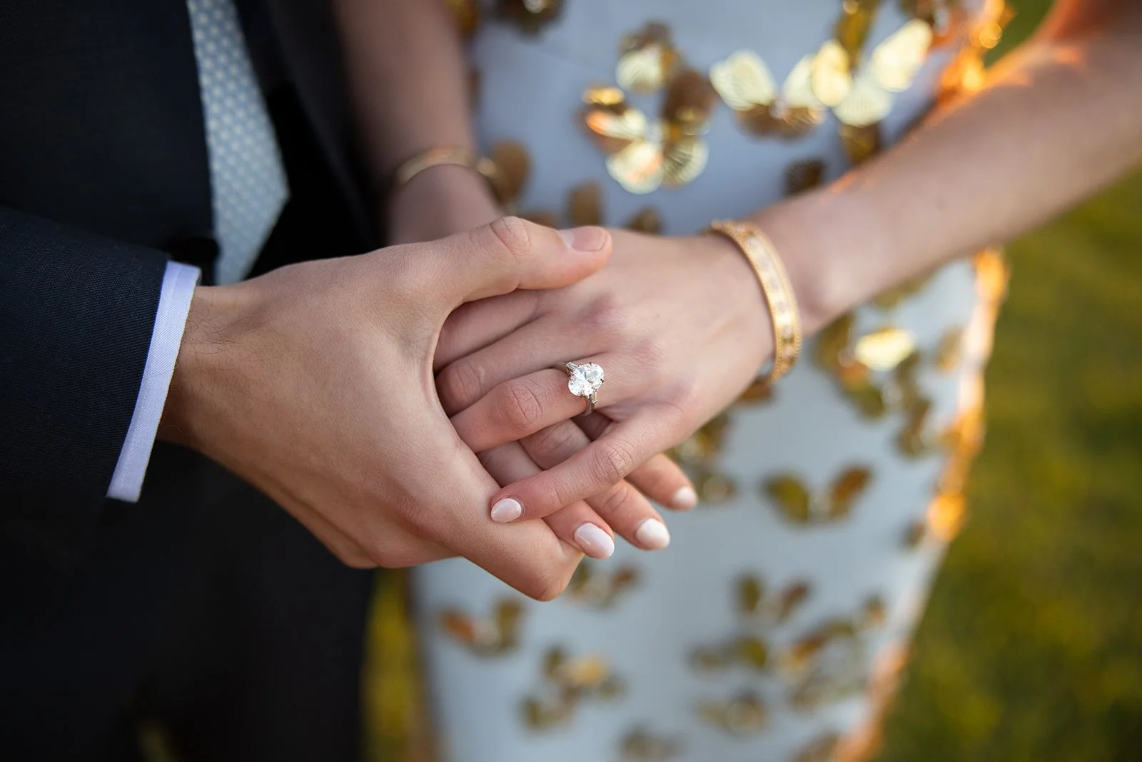 Close-up of a bride and groom holding hands, showing the bride's engagement ring and wedding band. The bride wears a gold bracelet, and the groom is dressed in a dark suit with a white shirt and tie. The background features a reflective surface with 