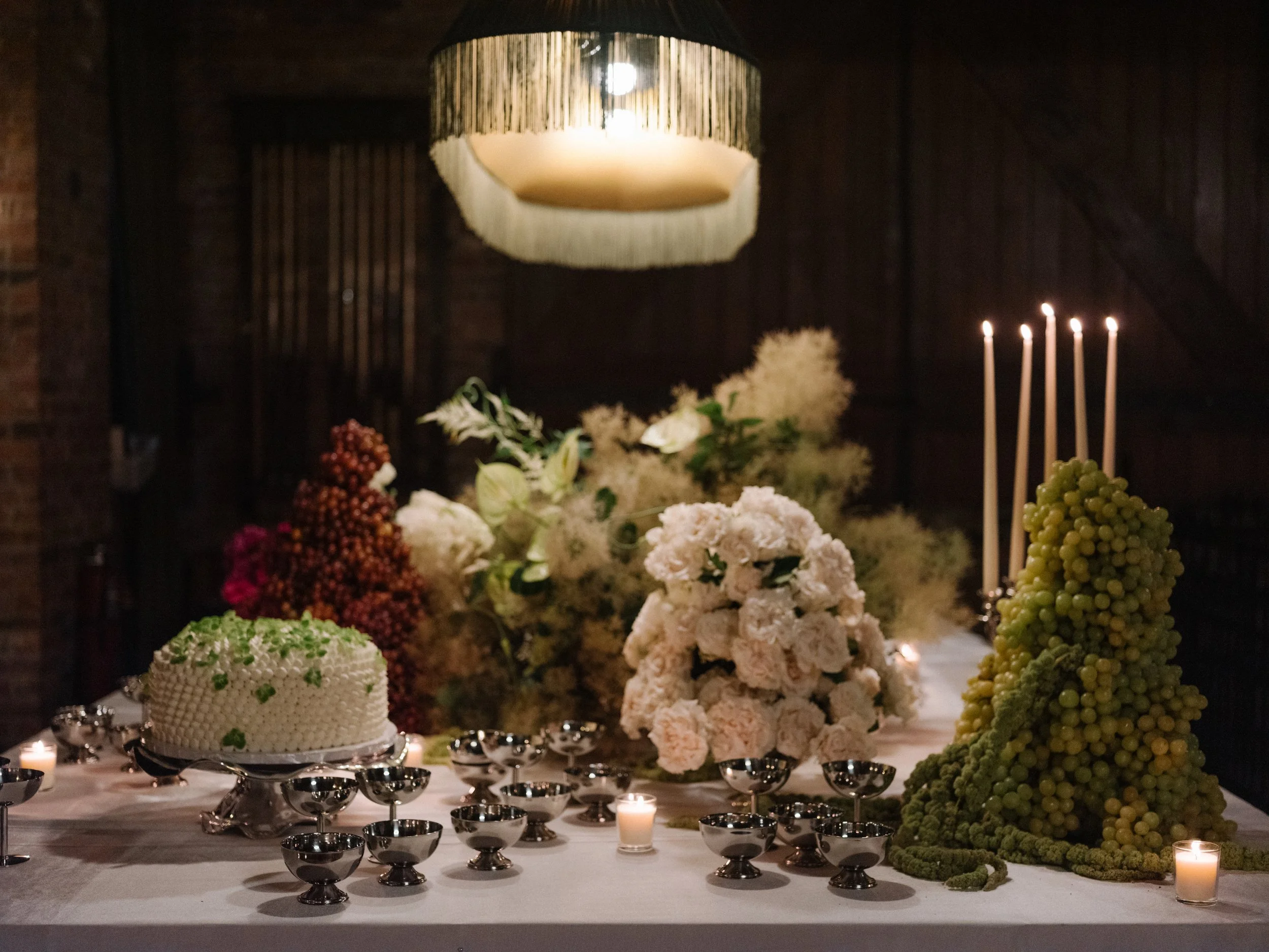 Table decorated with a white tablecloth, a white frosted cake with green decoration, large floral arrangements with white, pink, and red flowers, and green grapes shaped like trees, surrounded by lit candles, in a dimly lit room with dark wooden walls.