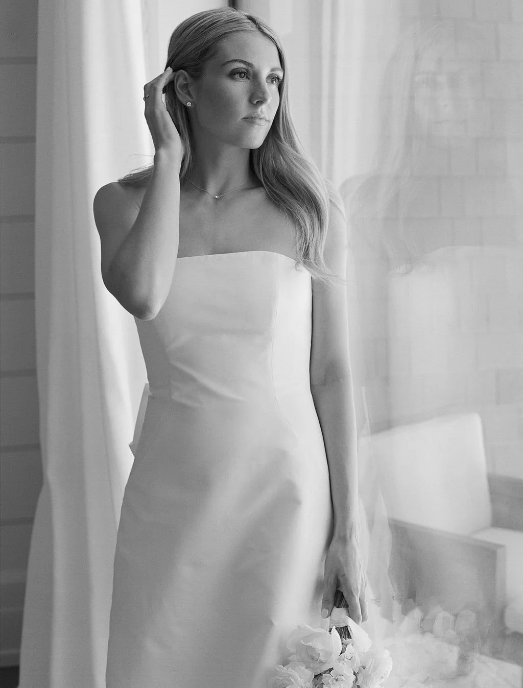 A woman in a strapless wedding dress is looking out a window while holding a bouquet of flowers, with a reflection visible on the glass.