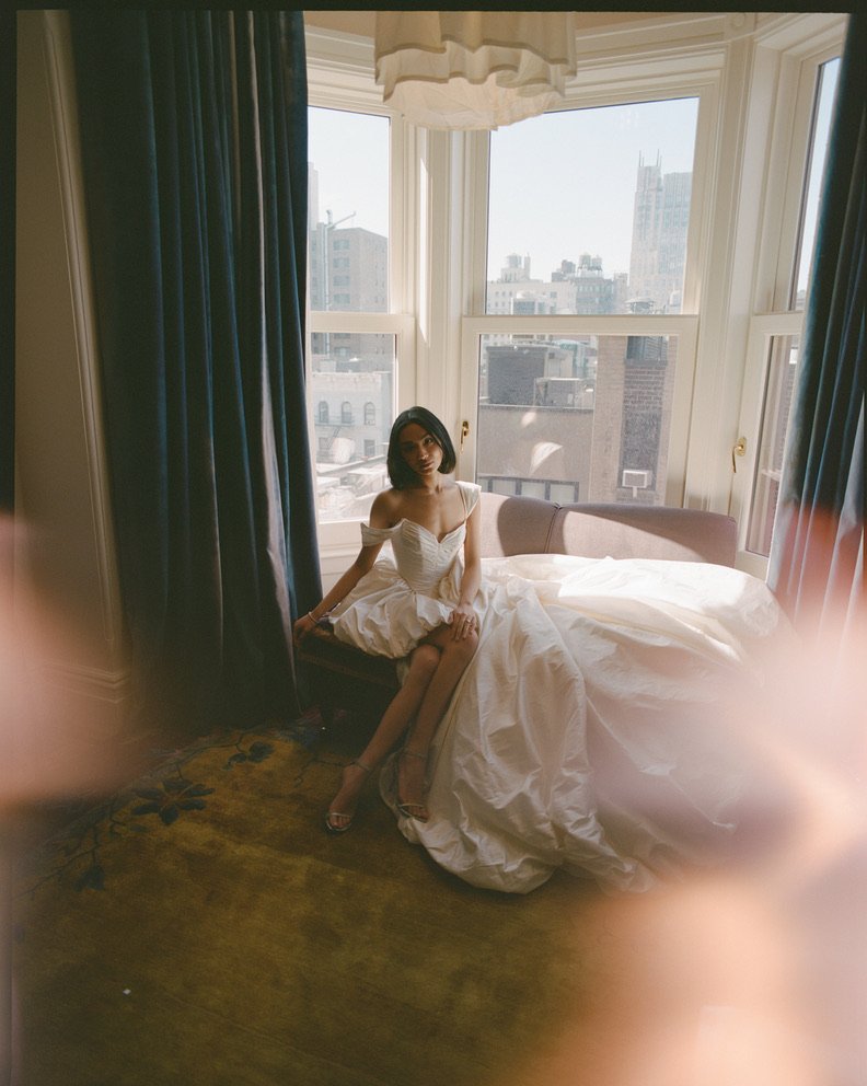 A woman in a white dress sitting on a bed near a large window with cityscape views, curtains on either side of the window, and sunlight illuminating the room.