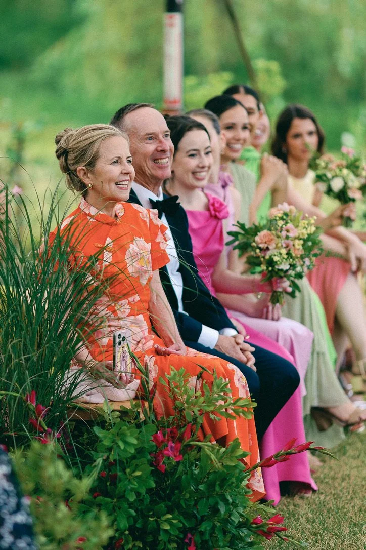 Group of people sitting outdoors, dressed in colorful formal attire, smiling, with women holding bouquets of flowers.
