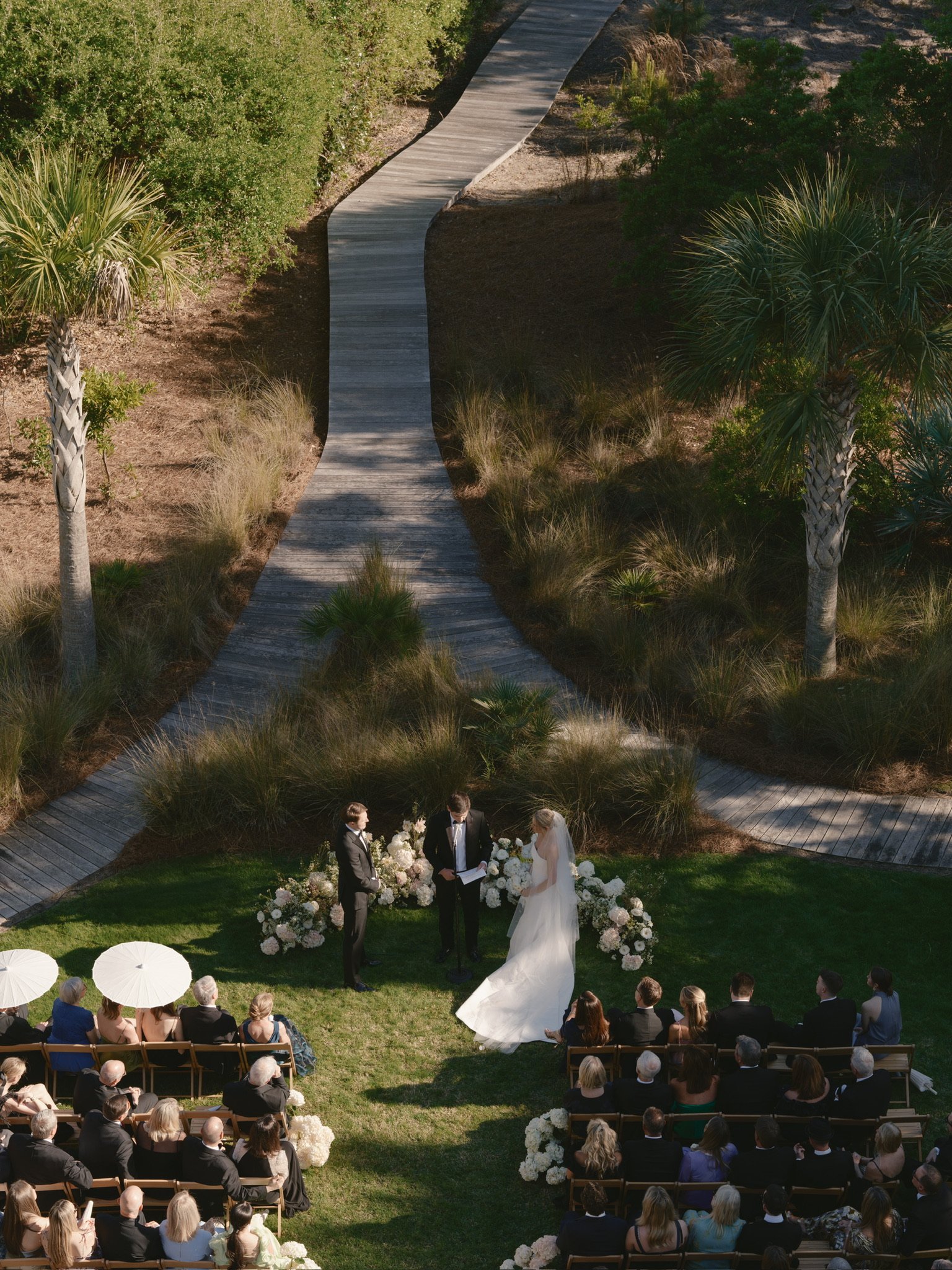 A wedding ceremony outdoors with a bride in a white dress and veil, groom in a tuxedo, and officiant standing under floral arrangements. Guests seated on benches, some holding umbrellas, watching on a grassy area surrounded by trees and a winding wooden pathway.