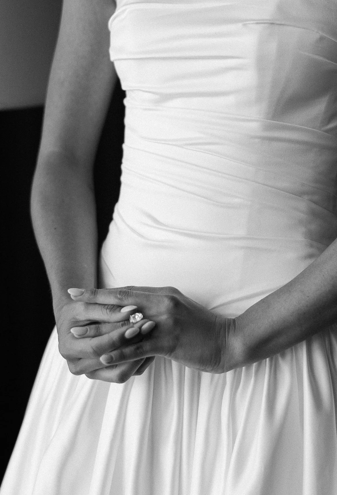 Close-up of a woman wearing a white dress, showing her hands with an engagement ring, in black and white.