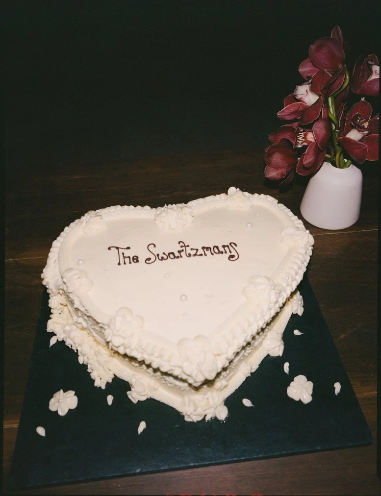 Heart-shaped white frosted cake with decorated border and the words 'The Swartzmans' written on top, placed on a black cake board with flower-shaped decorations, next to a white vase with dark red flowers.