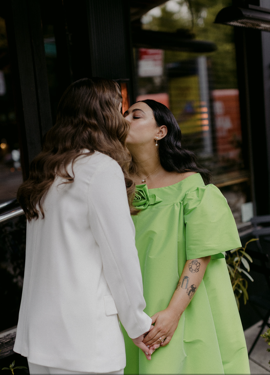 Two women kissing, one in a white suit and the other in a lime green dress, holding hands outside a building.