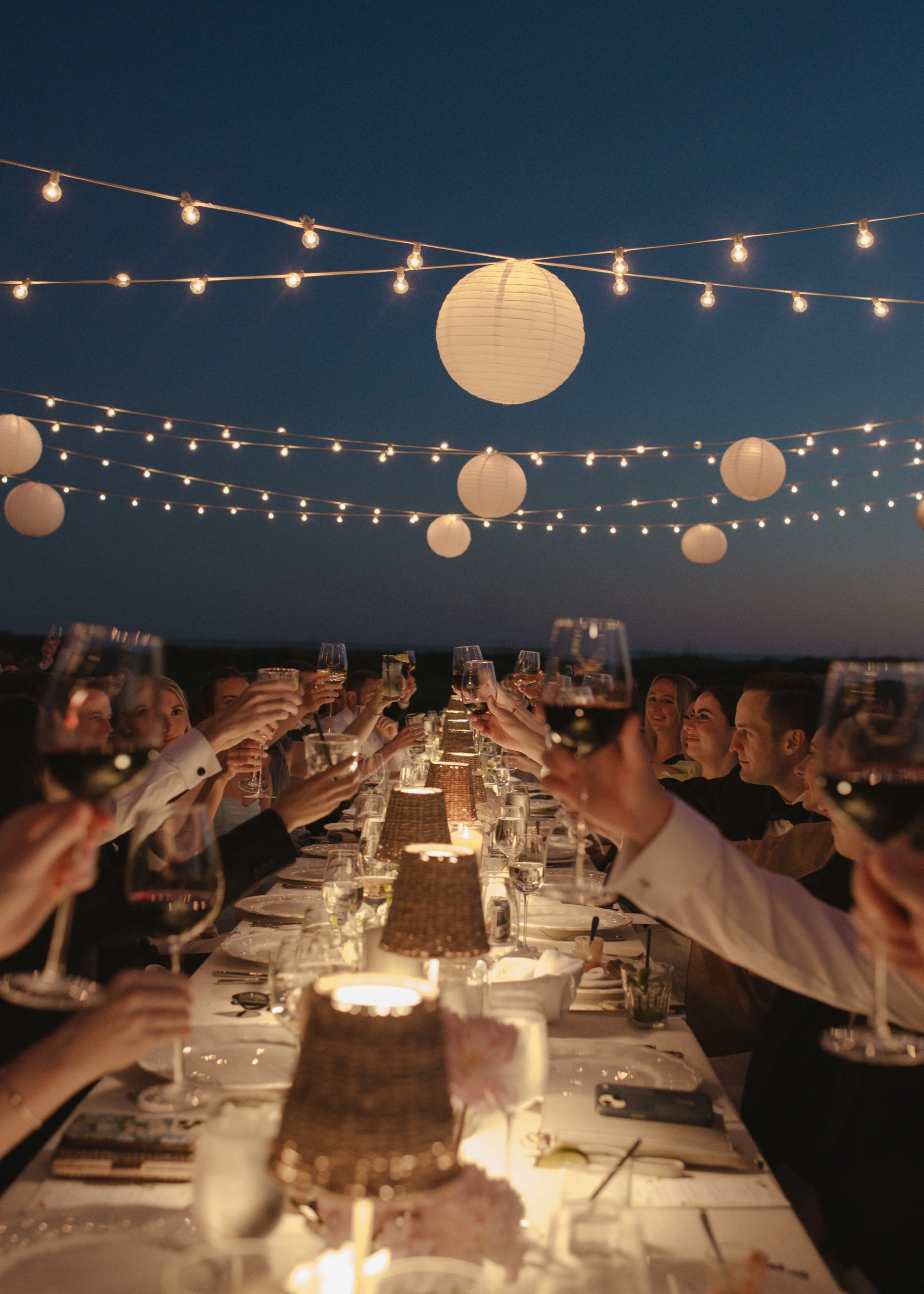 People raising glasses of wine at an outdoor evening dinner under string lights and paper lanterns.