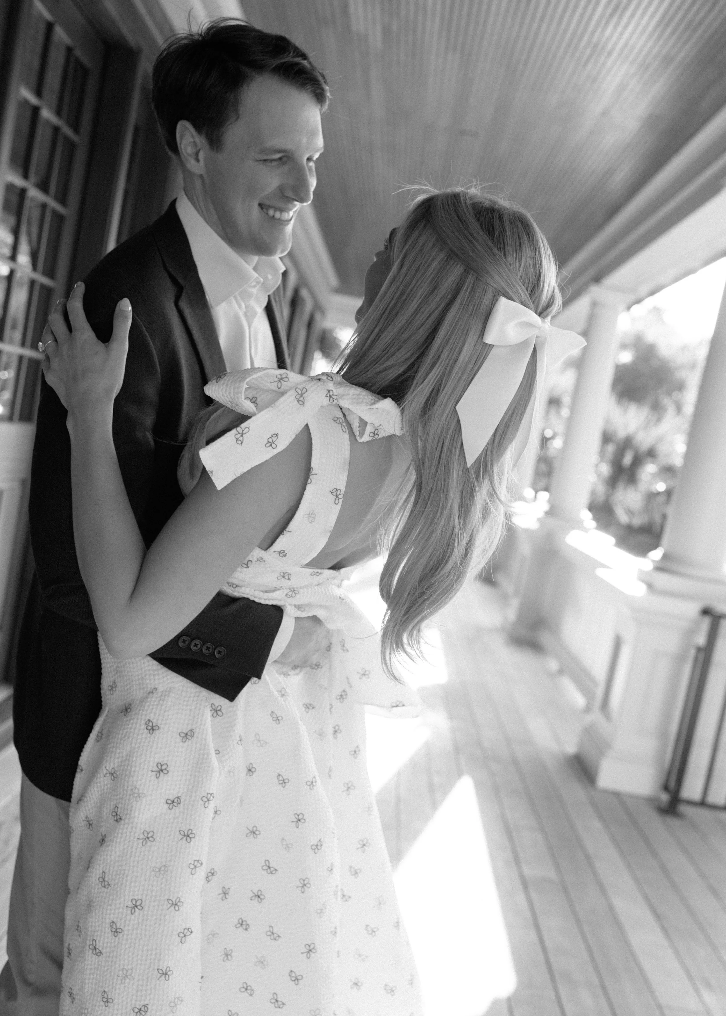 A black-and-white photo of a smiling man and woman on a porch; the man wears a suit jacket and white shirt, and the woman is in a dress with bows on the shoulders, with a bow in her hair.