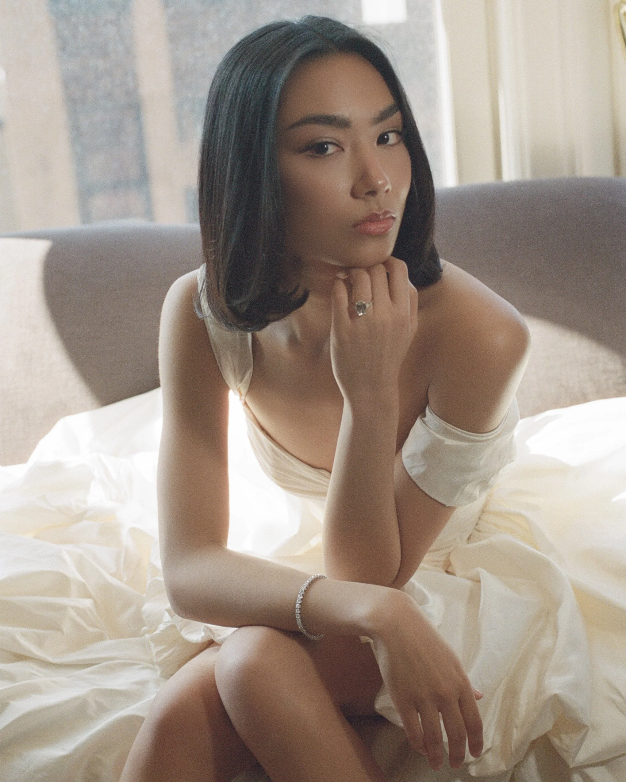 A woman with shoulder-length black hair sitting on a bed in a softly lit room, wearing a cream-colored dress with off-the-shoulder sleeves, and displaying rings and a bracelet.