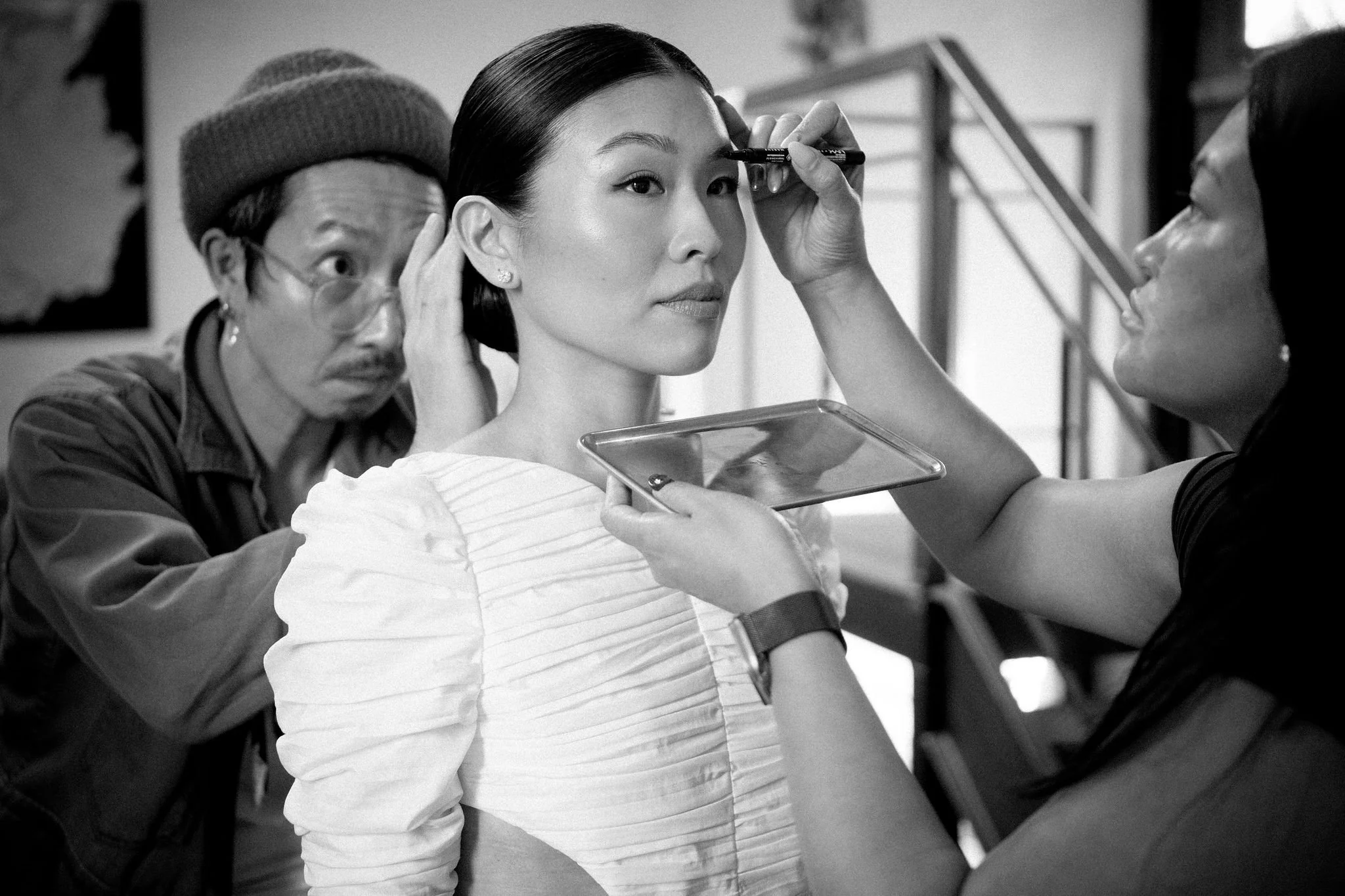 A woman with dark hair is getting her makeup done by a makeup artist, while an older man looks on, in a studio setting.
