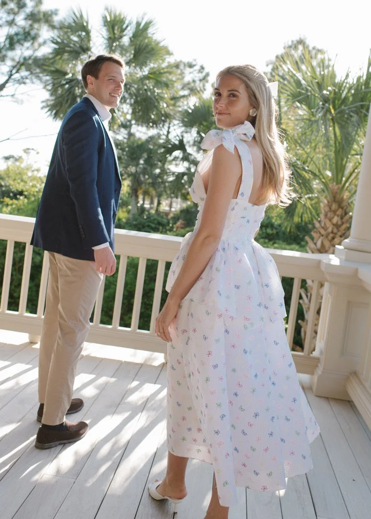 A young woman in a white, floral dress with bow straps standing on a porch, looking over her shoulder at a man in a navy blazer and beige pants, with palm trees in the background.