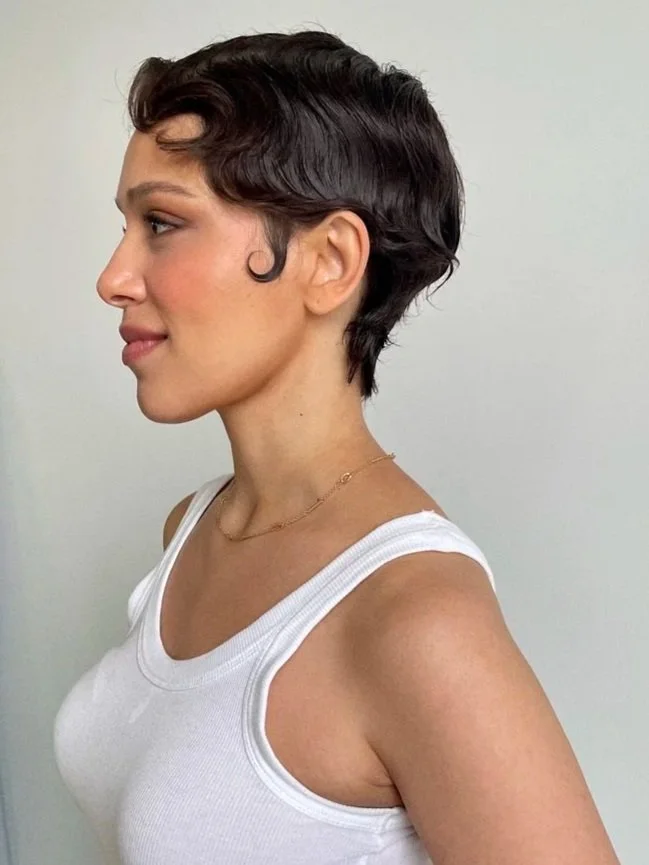 Side profile of a woman with short, dark hair, wearing a white tank top and a delicate gold necklace, standing against a plain light-colored background.