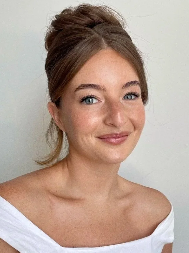 A young woman with light skin and reddish-brown hair styled in an updo, smiling softly, wearing a white off-shoulder top, standing against a plain light-colored background.