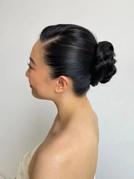 Side profile of a woman with black hair styled in an elegant updo bun, wearing a strapless top, against a plain background.