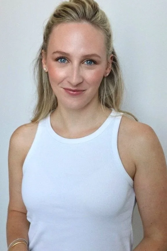 A woman with blonde hair and blue eyes smiling, wearing a white sleeveless top, standing in front of a plain light-colored wall.