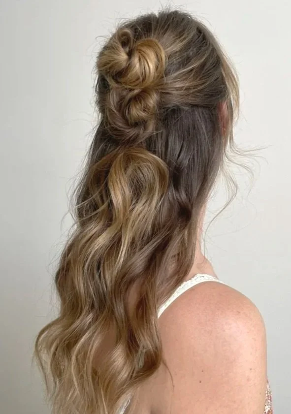 A woman with long, wavy light brown hair styled with a small bun on top and loose waves flowing down her back, wearing a white sleeveless top.
