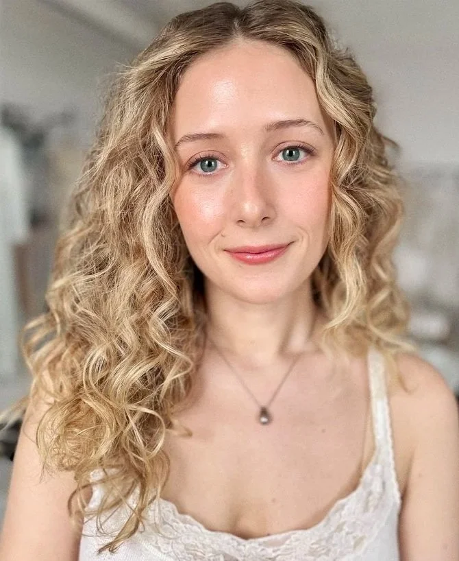 A young woman with long, curly blonde hair and blue eyes, smiling softly at the camera, wearing a white lace top and a delicate necklace, in a softly lit indoor setting.