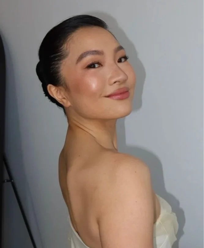 A woman with dark hair styled in an elegant updo, smiling softly, wearing natural makeup, and a strapless white dress, standing against a plain background.