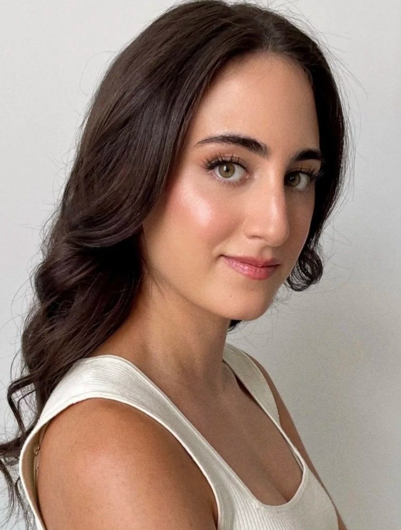 A young woman with long, wavy brown hair and light skin is posing for a portrait against a plain white background. She is wearing a light-colored sleeveless top and smiling softly.
