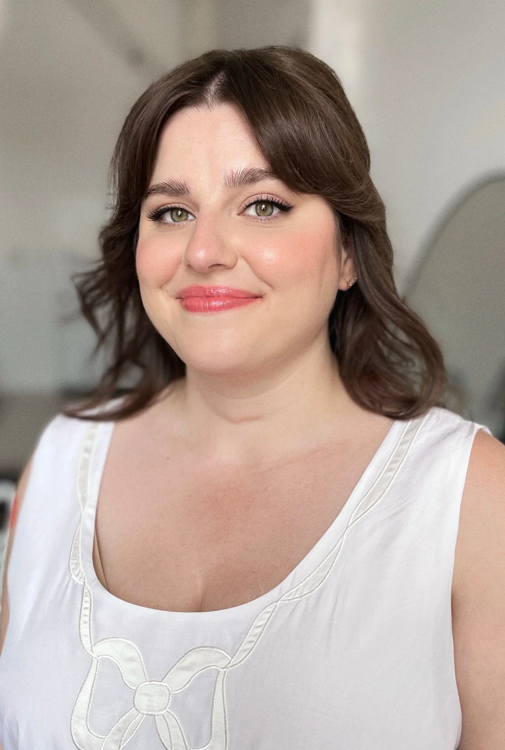 A woman with brown wavy hair, fair skin, wearing a sleeveless white top with decorative stitching, smiling softly in an indoor setting.