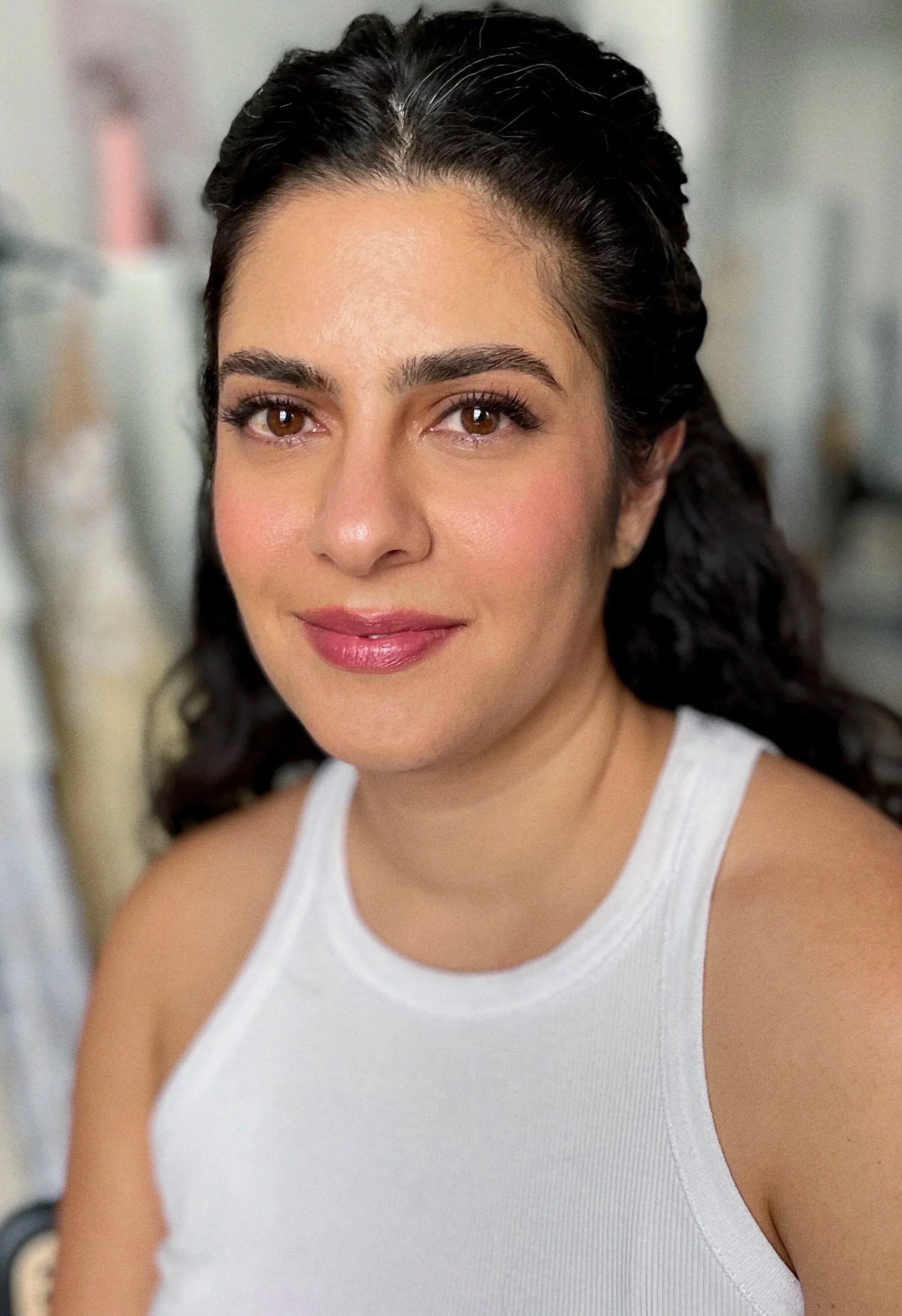 A woman with dark curly hair and brown eyes smiling and wearing a white sleeveless top.