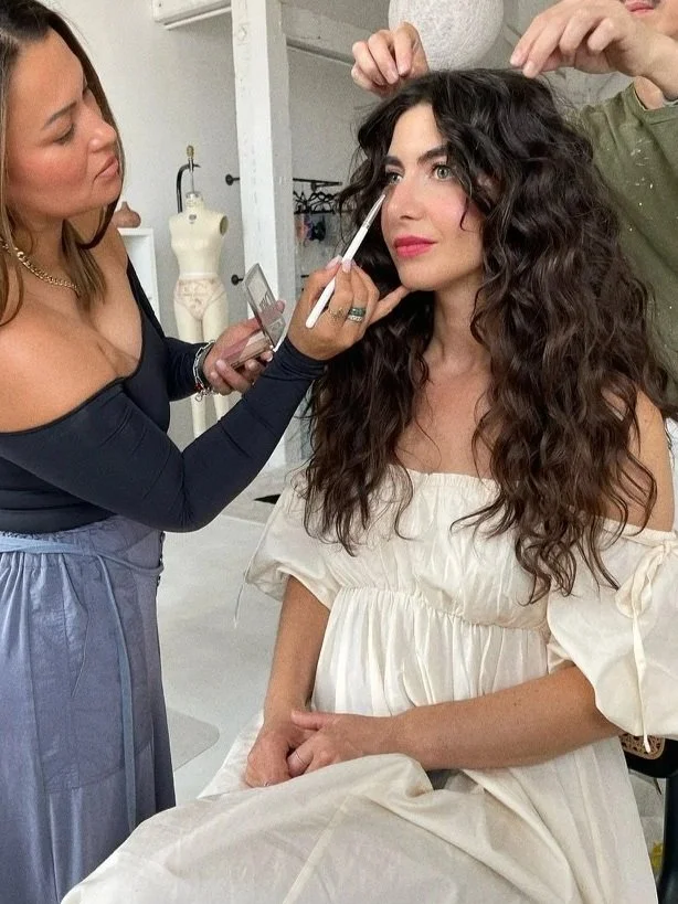 A woman with long, curly brown hair sits in a makeup chair while a makeup artist applies makeup to her face. The makeup artist holds a brush and is holding a mirror in her other hand. Someone else is adjusting the woman's hair, which is parted to the